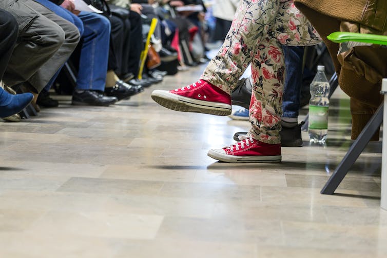Pies de muchas personas sentadas en una sala de espera, con alguien con zapatillas rojas y jeans floreados en primer plano y muchos con zapatos marrones y negros en el fondo.